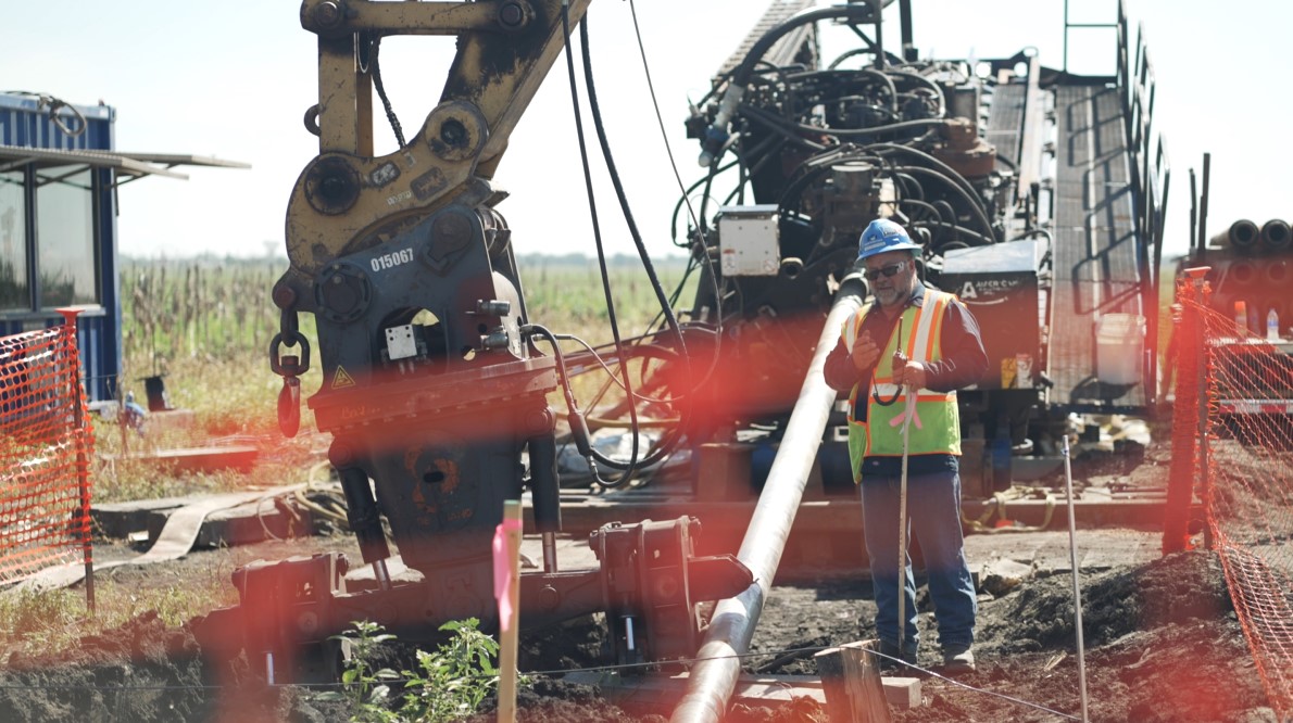 Construction site featuring a worker in safety gear overseeing Horizontal Directional Drilling (HDD) operations for oil and gas pipeline installation, with heavy machinery and equipment in the background.