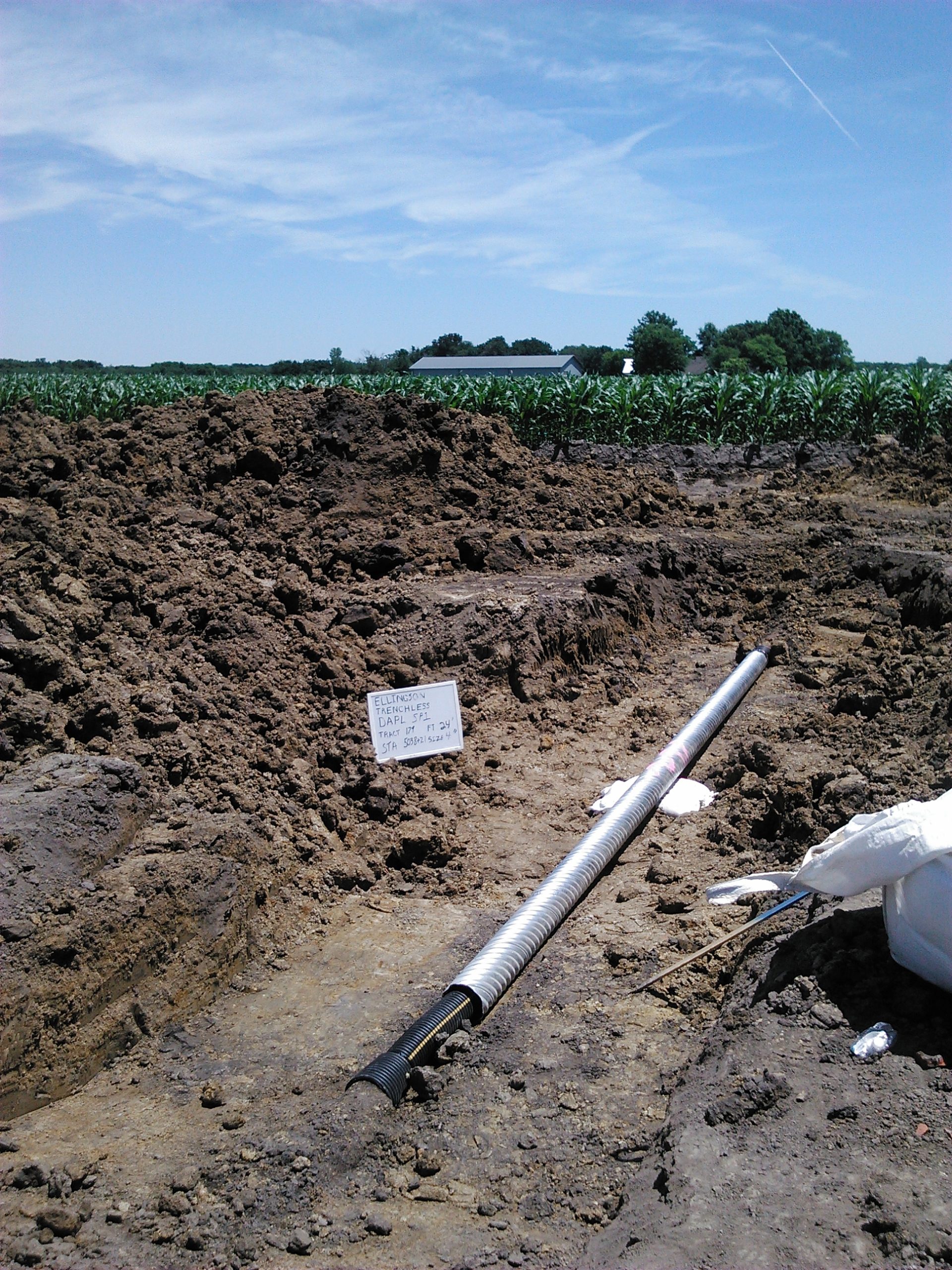 Excavated area showing drain tile installation with visible sign labeled "Ellingson Telemachine Darl ST", surrounded by agricultural land and crops, illustrating ECI Contracting's drain tile repair services.