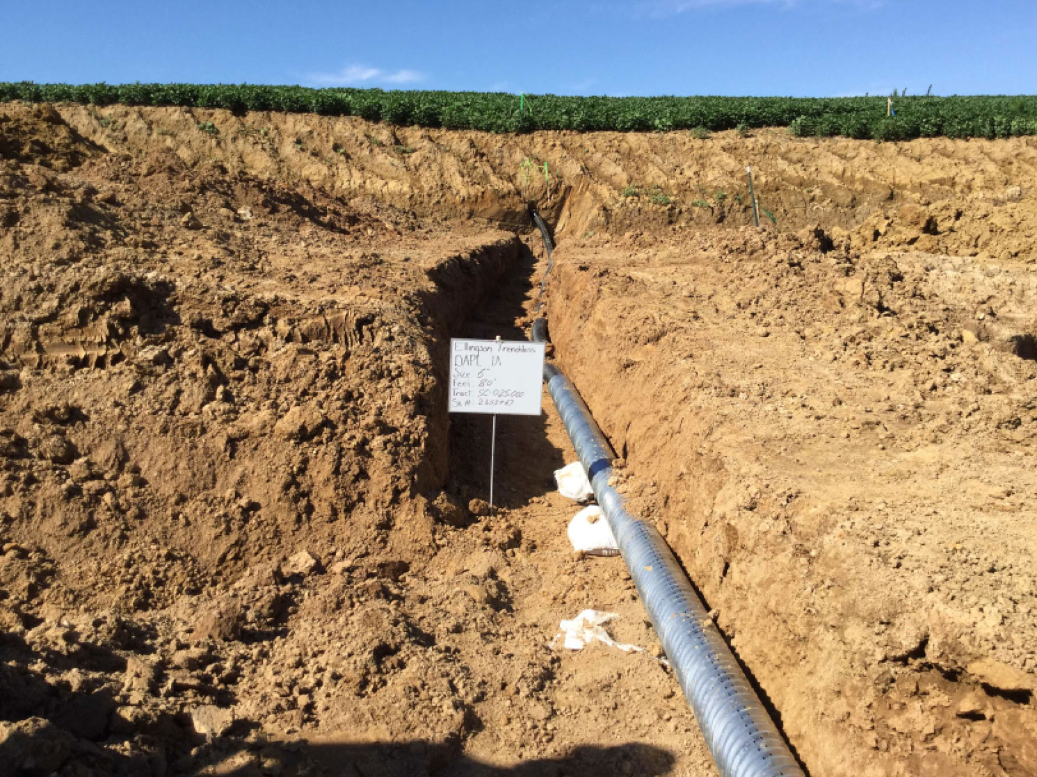 Drain tile installation site showing excavated trench with visible drainage pipe, sign indicating project details, and agricultural landscape in background, relevant to ECI's drain tile repair services for agricultural productivity.