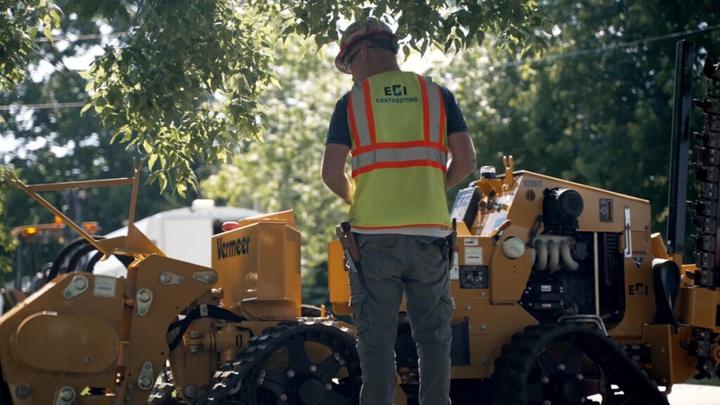 Worker operating Vermeer trenchless construction equipment, wearing ECI Contracting safety gear, in a green outdoor setting.